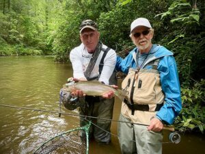 Two men stand in shallow water of a river showing a fish and a fly-fishing rod.